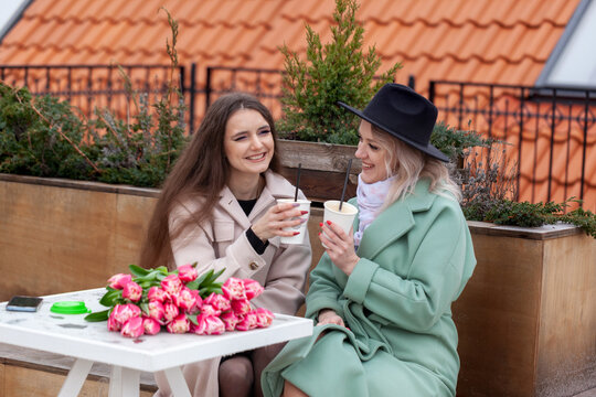 Two women enjoying coffee together outdoors, surrounded by blooming flowers, dressed in stylish coats, sharing laughter and conversation in a cozy urban setting
