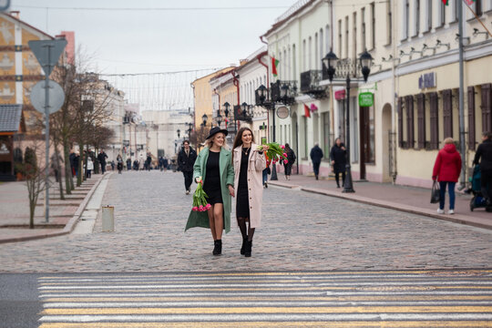 Two young women walk hand in hand across a city crosswalk. They wear stylish coats and hold flowers. The urban street is lively with pedestrians and shops.