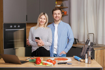 Smiling loving couple cooking salad together while standing on a kitchen at home and using mobile phone. Cute couple man and woman taking photo of food and taking pictures together while cooking.