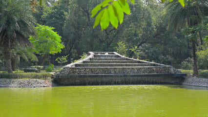 Stone pyramid structure stands near a pond with green water, surrounded by lush trees in a park-like setting. 