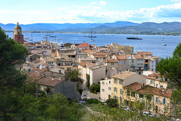 Obraz premium A panoramic view of Saint-Tropez, France, showing its pastel houses, church tower, and yachts anchored in the Mediterranean Sea against a mountain backdrop