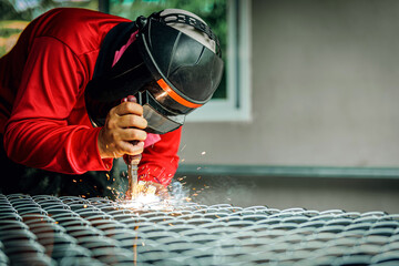 Welder wearing a red long-sleeved shirt and a protective mask is welding a steel grating, Industrial Welder With Torch
