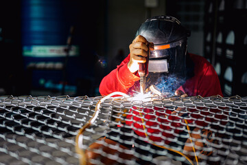 Welder wearing a red long-sleeved shirt and a protective mask is welding a steel grating,...