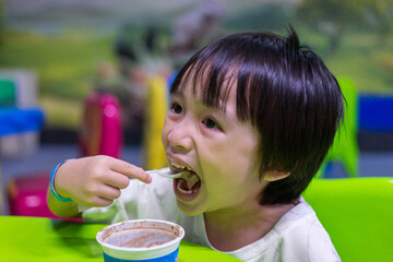The boy used a spoon to scoop up the ice cream in the cup and eat it deliciously. Kid wearing a white shirt and sitting on a chair scooping chocolate ice cream from a container.