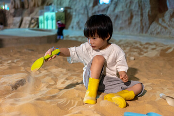 Kid happy face playing in the sandbox at the playground indoors. Cute boy plays with toys in the sand on the playground. Preschool creative activities and child development healthy.