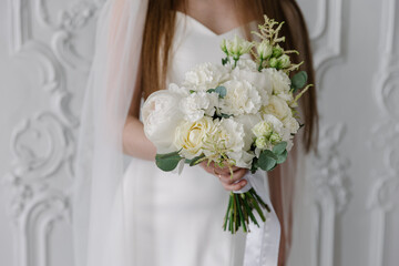 Elegant bride in a white satin gown holding a luxurious mixed floral bouquet featuring white peonies, cream roses, and eucalyptus against a classic stucco wall