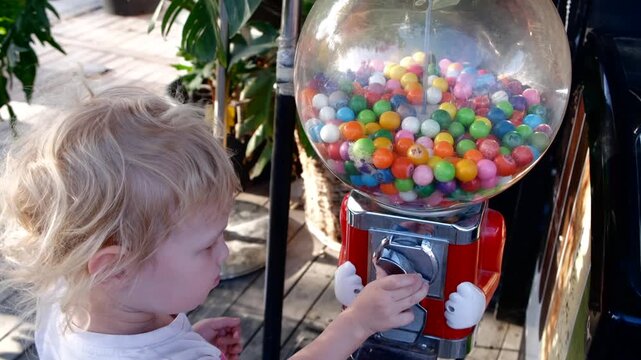 Curious toddler turning the handle of a colorful gumball machine. Bright candy balls in a transparent globe evoke childhood joy and sweet anticipation