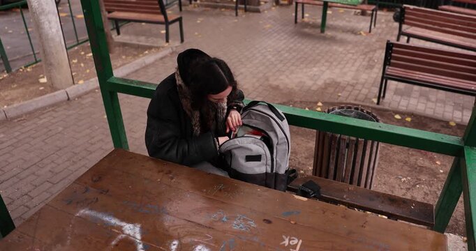 Woman student opens backpack on bench searching for notebook in city park. Teenager digs through pockets and finally finds diary smiling with relief