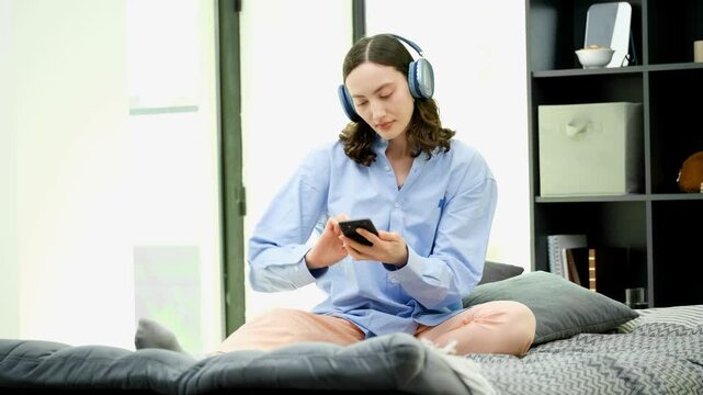 Young woman sits cross legged on sofa wearing headphones. Listener selects playlist on smartphone and nods head enjoying long music session at home