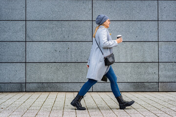Mature woman walking against background of gray modern wall in autumn. Copy space. Side view