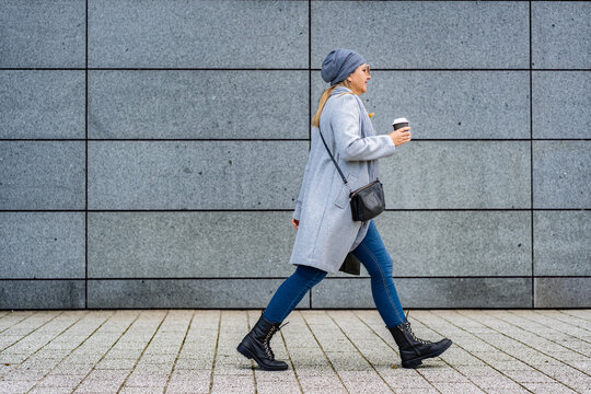 Mature woman walking against background of gray modern wall in autumn. Copy space. Side view - Powered by Adobe