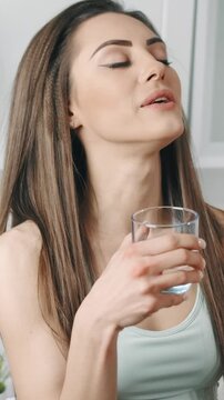 A woman stands in her kitchen, taking a moment to drink water from a clear glass. She looks content as she sips, enjoying hydration in a bright, cozy space.