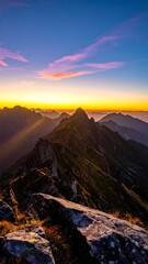 Mountain peaks bathed in sunrise hues.  Vast, jagged mountain ridges stretching into a colorful sunrise sky.  Rocky, foreground outcrop