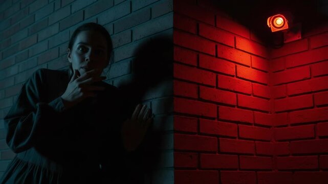 Frightened woman next to brick wall under security camera with an apprehensive gaze. Frightened woman stands in shadows, near brick wall corner and ominous red light.