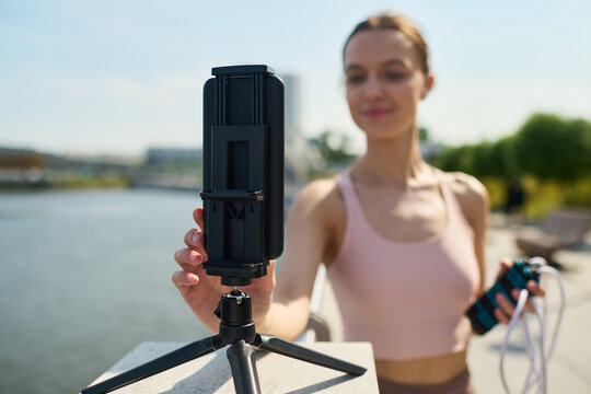 Young woman adjusting smartphone tripod outdoors near water holding jump rope in other hand preparing for fitness recording or workout in urban park