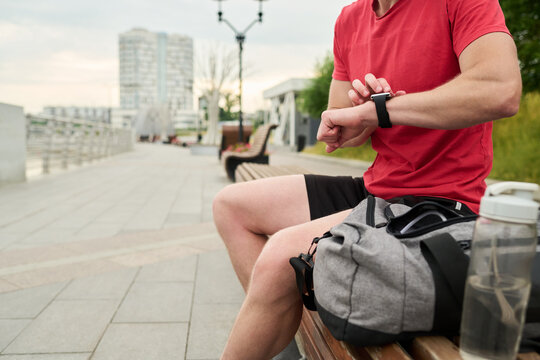 Young man sitting on bench checking smartwatch outdoors near sports bag and water bottle preparing for workout in urban park