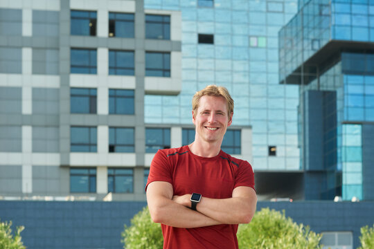Portrait of Young man smiling with arms crossed outdoors in front of modern glass building wearing smartwatch standing confidently looking at camera