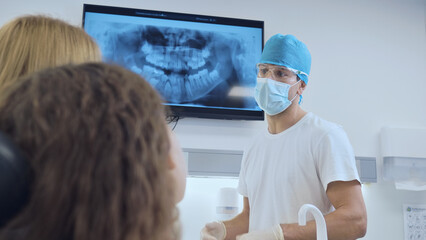 A dentist in a white uniform and blue cap explains dental X-ray results to a patient and observer...
