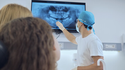 A dentist in a white uniform and blue cap explains dental X-rays to attentive patients in a modern clinic, emphasizing dental health education and patient care.