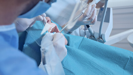 A dentist in a clinical setting performs a dental procedure, wearing gloves and using dental instruments. The patient, draped in a blue bib, sits in a dental chair.