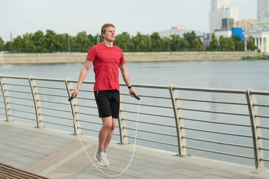 Young man exercising by jumping rope outdoors near river performing cardio workout on paved promenade with cityscape and trees in background focused expression