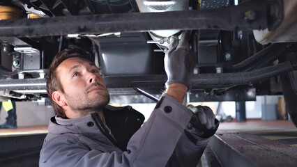 A focused mechanic works under a vehicle in a well-lit auto repair shop, using tools to inspect or repair the undercarriage, captured from an upward angle. His concentration highlights.