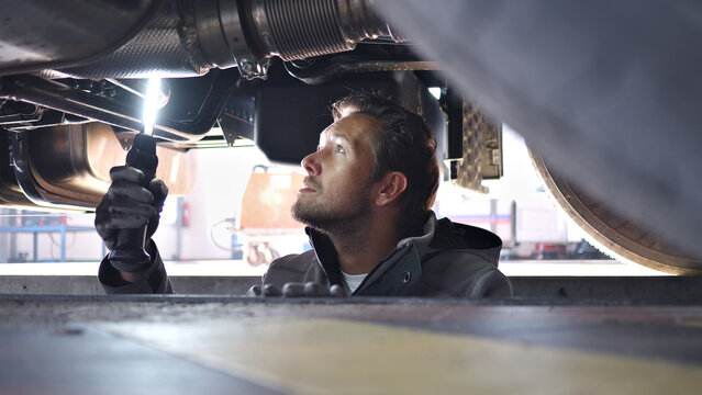 A focused mechanic uses a flashlight to inspect a car engine in a well-lit auto repair shop, highlighting the precision and expertise required in automotive maintenance, captured from below.