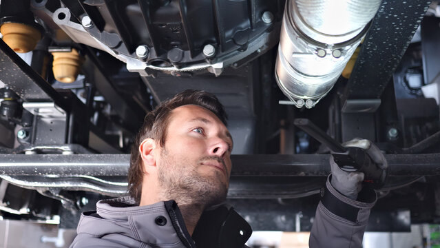 A focused mechanic inspects a vehicle's undercarriage in a well-lit auto repair shop, using a tool to examine components. The mechanic's attentive expression and professional attire highlight.