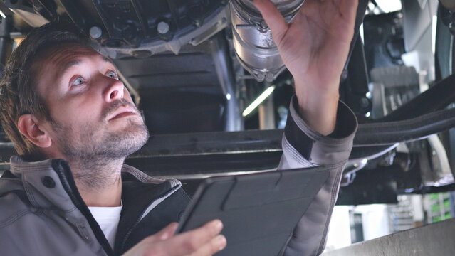 A mechanic examines a car's undercarriage with a tool and tablet in a well-lit auto shop, showcasing precision and technology in vehicle maintenance.