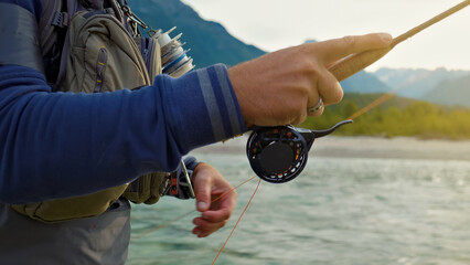 A focused fisherman in a blue shirt and vest casts a line into a serene freshwater lake, surrounded by majestic mountains. He skillfully handles a spinning reel, embodying the tranquility.