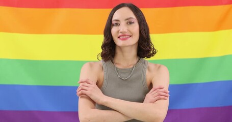 Happy young lesbian crosses arms and smiles on rainbow flag backdrop. Brunette queer lady radiates pride feeling in LGBT community slow motion