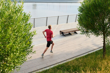 Young man jogging along riverside promenade running between green trees near water exercising outdoors athletic male maintaining fitness urban park setting