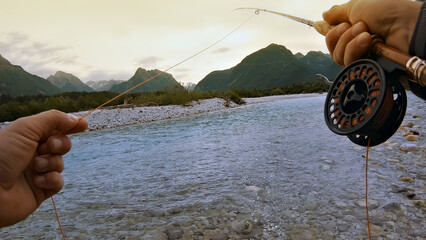 A person fly-fishing by a river, holding a fishing rod with a spinning reel. Majestic mountains...