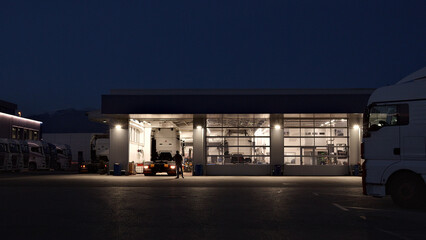 A lone mechanic works late in a well-lit auto repair shop, surrounded by parked trucks. The scene highlights the dedication and precision of a maintenance engineer.