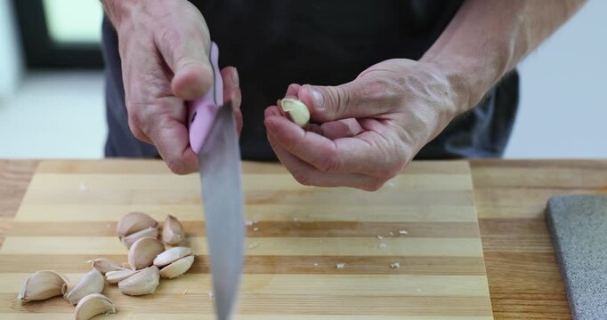 Man chef carefully peels garlic cloves using sharp knife at kitchen counter. Professional cook removes thin skin from garlic cloves. Slow motion