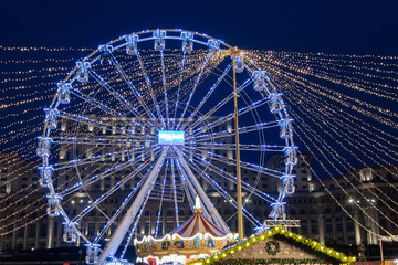 Illuminated ferris wheel with blue LED lights and carousel at Christmas market during evening. Festive holiday atmosphere with decorative string lights against dark blue sky