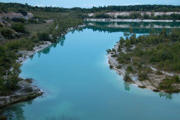A vibrant teal lake winds through a former quarry, surrounded by green trees and light-colored banks under a soft sky, reflecting the serene environment.