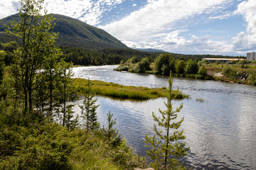 A winding river flows through a lush, forested valley beneath a partly cloudy sky, with a mountain in the distance and a large industrial structure on the far bank.