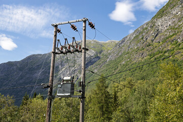 An electricity pole with a power transformer stands among green trees against a majestic mountain...