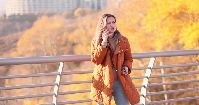 Woman in coat argues emotionally while talks on smartphone on bridge in autumn park. Angry lady has problematic conversation via cellphone outdoors