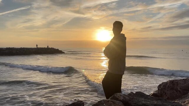 A man stands on a rock overlooking the ocean. The sky is a mix of orange and pink hues, and the sun is setting. The man is lost in thought, and the scene has a peaceful, serene atmosphere