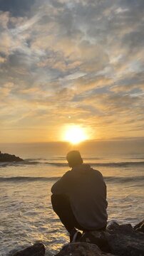 A man sits on a rock overlooking the ocean, watching the sun set. The sky is filled with clouds, creating a moody atmosphere. The man is lost in thought