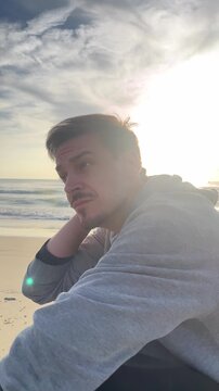 A man is sitting on the beach with his head down. He is wearing a gray sweatshirt and has a beard. The sky is cloudy and the sun is shining