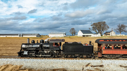 Fototapeta premium A vintage steam train moves along the tracks in a rural area. It is surrounded by golden fields and distant farm buildings under a dramatic cloudy sky. The scene captures a moment of nostalgia.