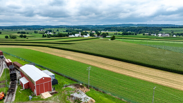 Expansive green fields stretch across the rural landscape of Pennsylvania, with farm structures visible in the foreground. Rolling hills and a cloudy sky create a serene farming scene.