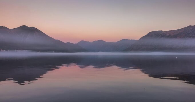 Misty dawn before sunrise over the calm Kotor Bay in Montenegro, with soft purple light, still water, and distant mountain silhouettes fading into morning fog. Time lapse