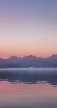 Misty dawn before sunrise over the calm Kotor Bay in Montenegro, with soft purple light, still water, and distant mountain silhouettes fading into morning fog. Time lapse