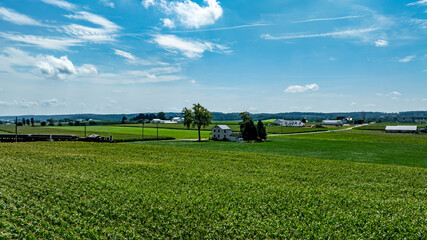 Lush green fields stretch across the landscape under a clear blue sky with fluffy clouds. A small...