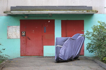 A motorcycle covered with a blue cover stands near the entrance to an apartment building, Dal'nevostochny Prospekt, Saint Petersburg, Russia, October 07, 2025