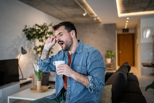 Tired man yawning, rubbing eye, drinking morning coffee